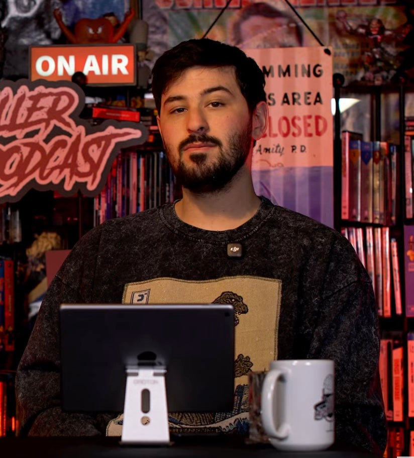 Man sitting at a desk with a tablet and mug in a room with various signs and decor.