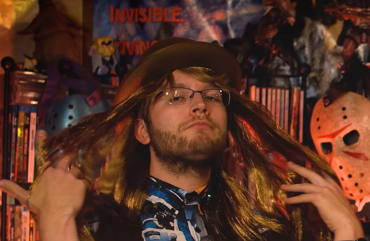 Person with long hair and beard wearing glasses and a hat, sitting at a table with a laptop in a room with Halloween decorations.