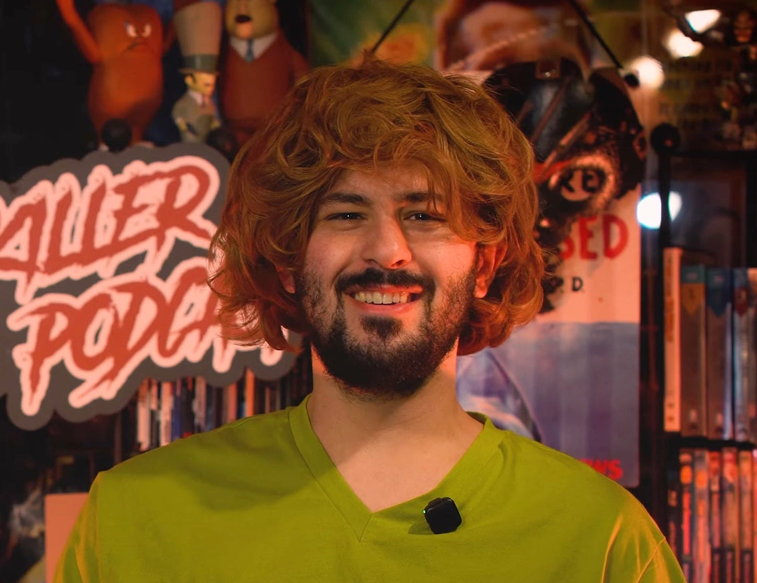 Person in a green shirt sitting at a desk with a laptop, surrounded by bookshelves and posters.
