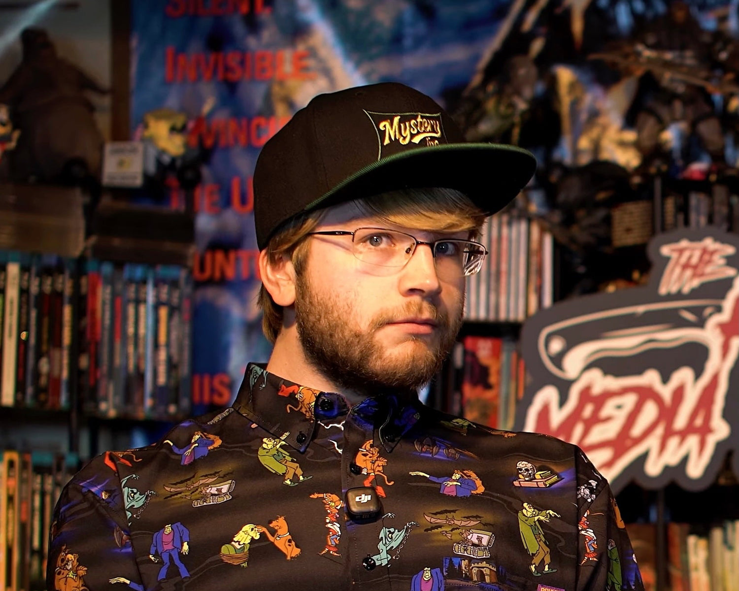 Man wearing a patterned shirt and cap in a room with bookshelves and posters.
