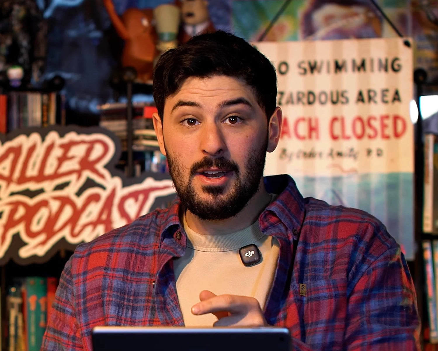Man in plaid shirt sitting at a desk with a laptop and mug, in front of a colorful background with signs.